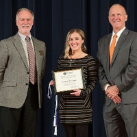 Award recipient in a black, grey, and white striped sweater dress posing for a photo on stage with Doctor Potteiger and Mark Luttenton